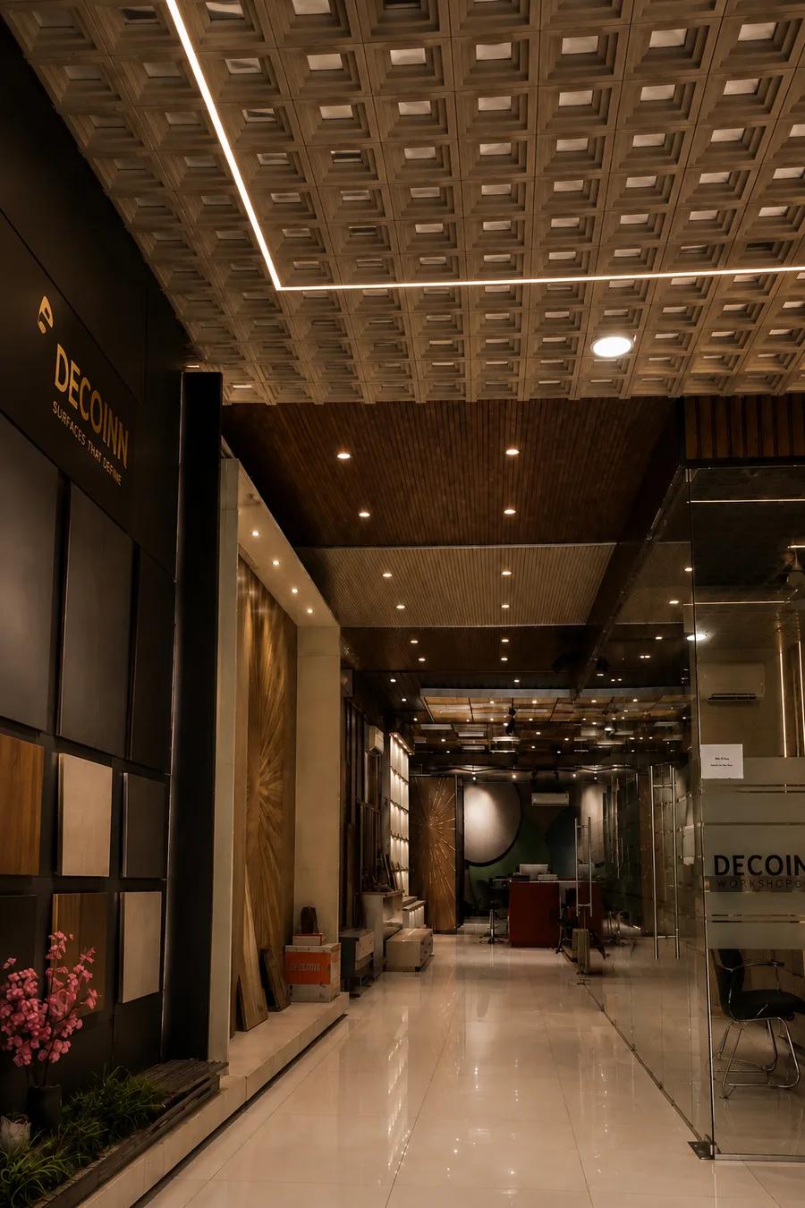 The DECOINN display gallery in Ludhiana: a long, softly lit corridor with a polished stone floor. The black feature wall on the left carries the gold DECOINN mark above its line SURFACES THAT DEFINE, with square sample panels in wood and stone finishes set into the wall beneath it. A deep coffered ceiling runs overhead and gives way to a fluted timber soffit, and a glazed partition down the right, lettered DECOINN Workshop, carries the eye to a desk at the far end.